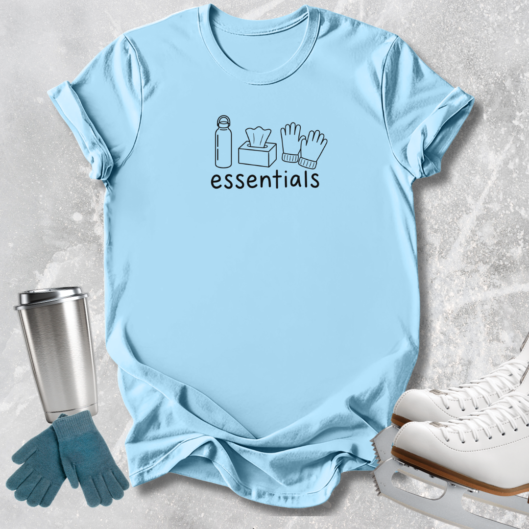 A light blue graphic t-shirt laid flat on a textured gray background, featuring a black line art design of winter essentials (water bottle, tissues, gloves, skates) with the word 'essentials' printed below. A stainless steel travel mug, blue gloves, and white ice skates are styled around the shirt.