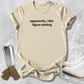 A beige graphic t-shirt with the text 'apparently, i like figure skating' printed in black. The t-shirt is styled in a flat-lay arrangement on a textured gray background, accompanied by ice skates, gloves, and a travel tumbler.