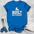 A bright blue short-sleeve T-shirt laid flat on a light gray textured surface, printed with a white ice skate graphic and the words 'BUILT DIFFERENT.' Blue gloves and white ice skates are placed around it as styling props.