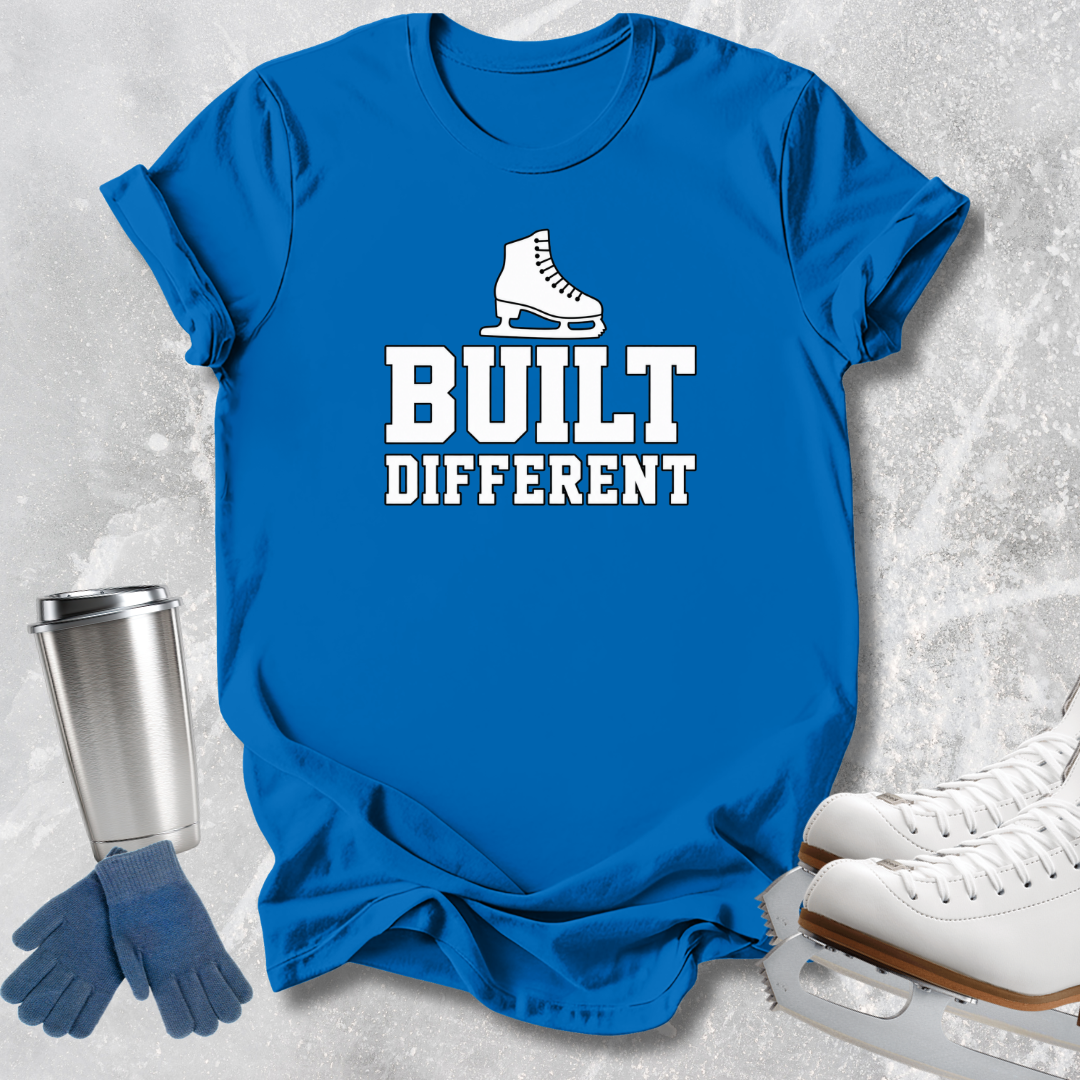 A bright blue short-sleeve T-shirt laid flat on a light gray textured surface, printed with a white ice skate graphic and the words 'BUILT DIFFERENT.' Blue gloves and white ice skates are placed around it as styling props.