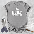 A heather gray graphic T-shirt laid flat on a light textured background, featuring a white ice skate icon above the words 'BUILT DIFFERENT' in bold white lettering. A stainless steel travel mug, gray gloves, and white ice skates are arranged around the shirt as styled props.