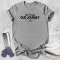 A heather gray short-sleeve t-shirt with the black printed text 'Her Dream OUR JOURNEY' and a small ice skate graphic, styled in a flat lay with white ice skates, gray gloves, and a stainless steel travel mug on a light gray textured background.