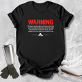 A black graphic t-shirt laid flat on a light gray textured background, styled with ice skates, gloves, and a metal cup. The shirt features a humorous warning label printed in red and white text.