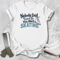 A white short-sleeve graphic T-shirt laid flat on a light gray textured surface, with the text 'Nobody Said There'd Be This Much SKATING' and a skater boot illustration. Ice skates, gloves, and a metal cup are arranged around the shirt as styled props.