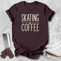 A dark brown short-sleeve t-shirt with the words 'SKATING & COFFEE' printed in cream on the front, styled in a flat lay with white ice skates, a stainless steel travel mug, and a pair of brown gloves on a light gray textured background.