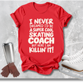 A red short-sleeve graphic T-shirt laid flat on a light gray textured surface, with white text reading 'I NEVER DREAMED I'D BE A SUPER COOL SKATING COACH BUT HERE I AM KILLIN' IT!'. Ice skates, gloves, and a metal cup are placed around it as styled props.