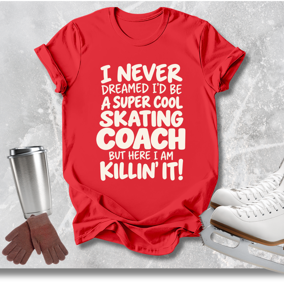 A red short-sleeve graphic T-shirt laid flat on a light gray textured surface, with white text reading 'I NEVER DREAMED I'D BE A SUPER COOL SKATING COACH BUT HERE I AM KILLIN' IT!'. Ice skates, gloves, and a metal cup are placed around it as styled props.