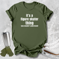 An olive green graphic t-shirt with the text 'it's a figure skater thing (you wouldn't understand)' in white, styled in a flat-lay with white ice skates, a silver travel mug, and green gloves on a textured gray background.
