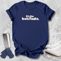 A navy blue short-sleeve t-shirt with the white printed text 'It's the Ice's Fault' and a small ice skate graphic, styled in a flat lay with ice skates, gloves, and a travel tumbler on a light gray textured background.
