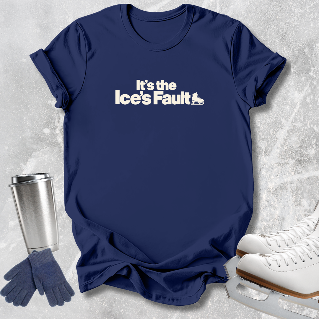 A navy blue short-sleeve t-shirt with the white printed text 'It's the Ice's Fault' and a small ice skate graphic, styled in a flat lay with ice skates, gloves, and a travel tumbler on a light gray textured background.