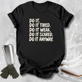 A black short-sleeve graphic T-shirt laid flat on a light textured surface, with cream text reading 'DO IT. DO IT TIRED.