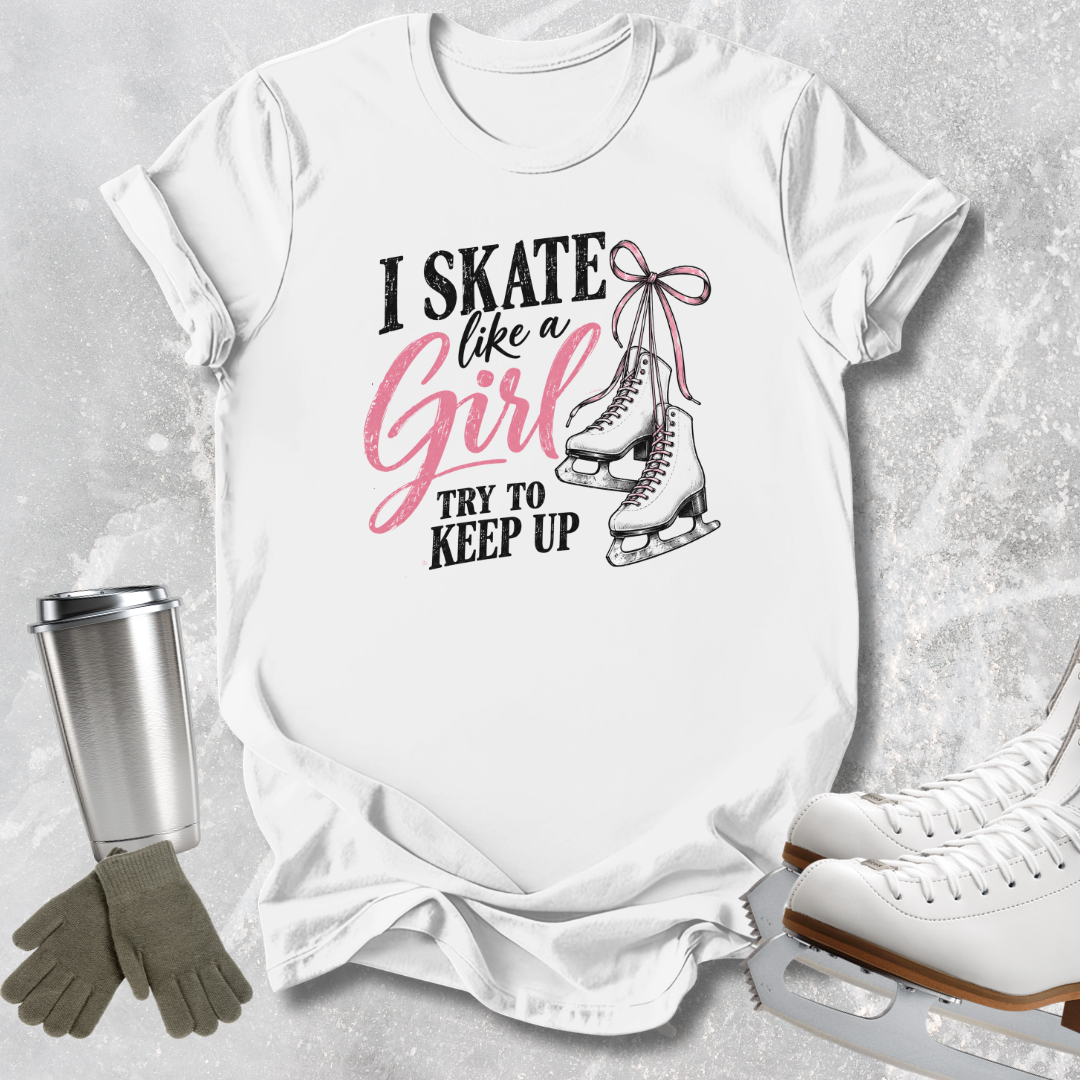A white graphic t-shirt laid flat on a light gray textured surface, featuring the text 'I SKATE like a Girl TRY TO KEEP UP' in black and pink script, alongside an illustration of ice skates. A stainless steel tumbler, gray gloves, and white ice skates are styled around the shirt.