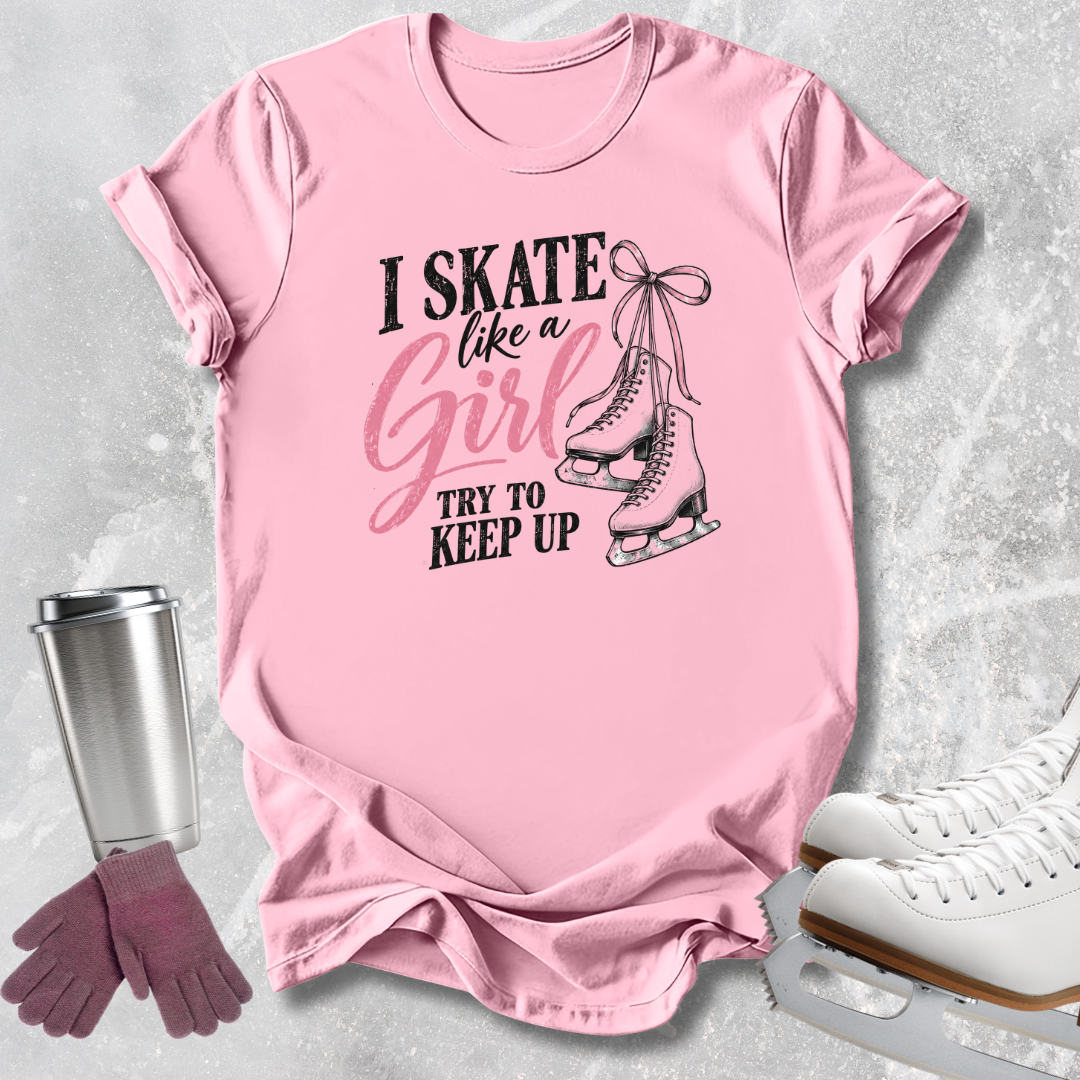 A pink short-sleeve graphic T-shirt laid flat on a light gray textured surface, with a silver travel mug, mauve gloves, and white ice skates around it. The shirt reads 'I SKATE like a Girl TRY TO KEEP UP' with a black-and-pink graphic of ice skates tied together.