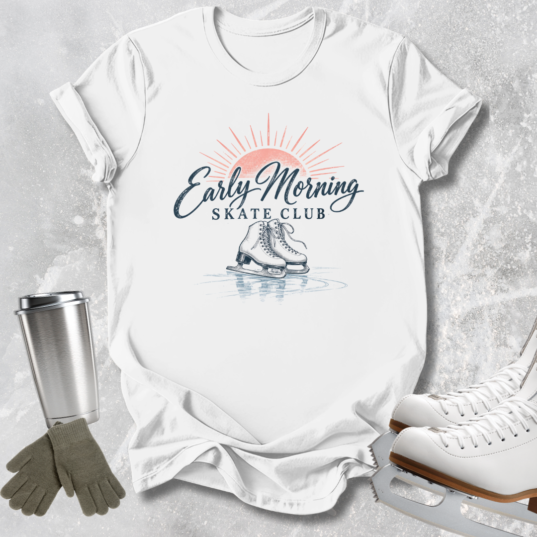 A white graphic T-shirt laid flat on a light gray textured surface, featuring a retro-style 'Early Morning Skate Club' print with a sun, skates, and rink reflection. A stainless steel cup, gray gloves, and white ice skates are arranged around the shirt as styled props.