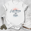 A white graphic T-shirt laid flat on a light gray textured surface, featuring a retro-style 'Early Morning Skate Club' print with a sun, skates, and rink reflection. A stainless steel cup, gray gloves, and white ice skates are arranged around the shirt as styled props.