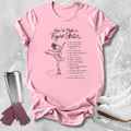 A pink graphic T-shirt laid flat on a light gray textured surface, featuring a black illustration of a figure skater and the text 'How to Make a Figure Skater' with a list of tips. White ice skates, a metal travel mug, and mauve gloves are arranged around the shirt as styling props.