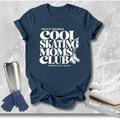 A navy blue graphic t-shirt with white text reading 'PROUD MEMBER COOL SKATING MOMS CLUB' and 'EMBRACING CHAOS', styled in a flat lay with ice skates, gloves, and a metal cup on a light gray textured background.