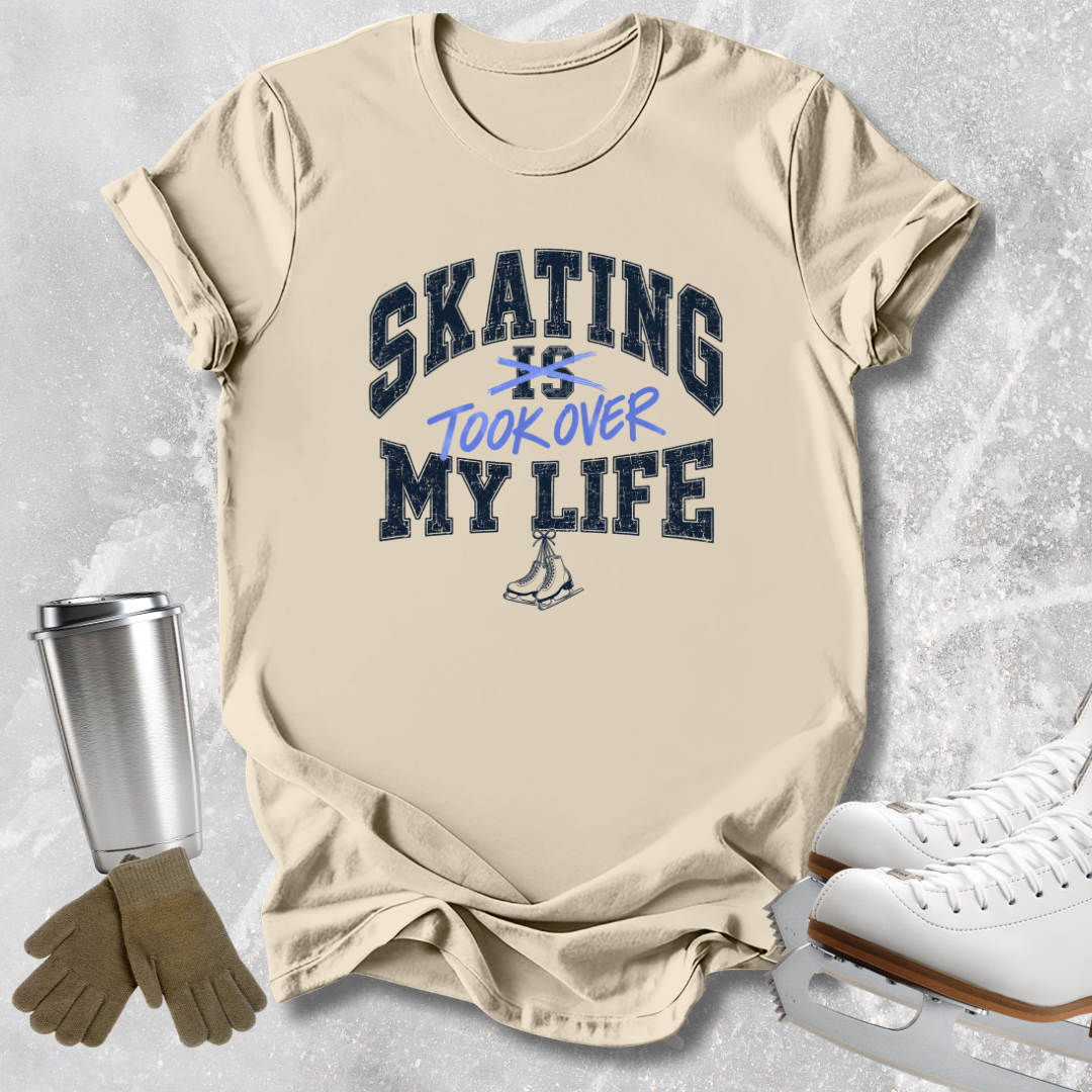 A beige graphic t-shirt laid flat on a light gray textured background, surrounded by ice skating props including white ice skates, a silver travel tumbler, and tan gloves. The t-shirt features the text 'SKATING IS TOOK OVER MY LIFE' in navy and blue lettering with a small skater graphic below.