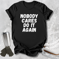 A black short-sleeve graphic T-shirt laid flat on a light gray textured surface, with large distressed white text reading 'NOBODY CARES DO IT AGAIN.' Dark gloves and a metal travel tumbler appear at left, and white ice skates appear at right as styled props.