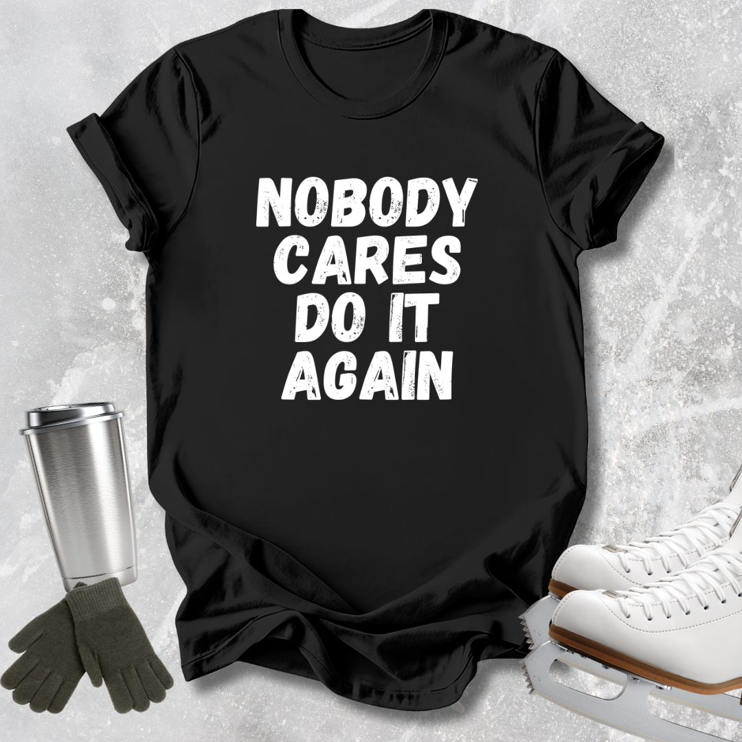 A black short-sleeve graphic T-shirt laid flat on a light gray textured surface, with large distressed white text reading 'NOBODY CARES DO IT AGAIN.' Dark gloves and a metal travel tumbler appear at left, and white ice skates appear at right as styled props.