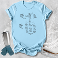 A light blue graphic t-shirt laid flat on a textured gray background, styled with ice skates, gloves, and a metal tumbler. The t-shirt features a black line drawing of a figure skater and text listing qualities like grace, toughness, confidence, and what it really takes.
