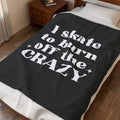 A dark charcoal gray blanket with bold white text reading 'I skate to burn off the CRAZY' is draped over a bed in a bedroom setting with a wooden nightstand and wood floor.