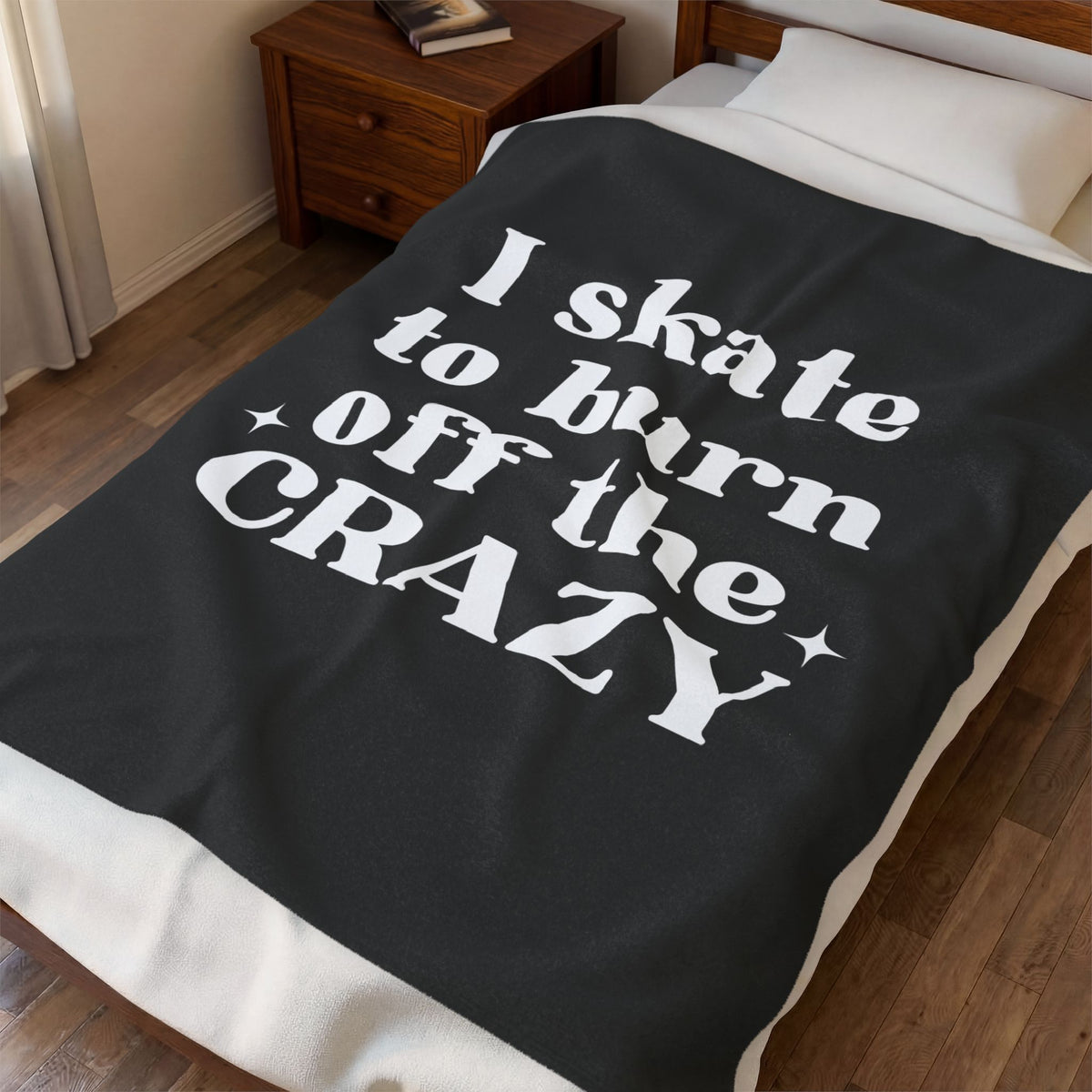 A dark charcoal gray blanket with bold white text reading 'I skate to burn off the CRAZY' is draped over a bed in a bedroom setting with a wooden nightstand and wood floor.
