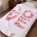 A light pink blanket with a large floral 'RO' design and a white ice skate motif, shown draped over a bed in a bedroom setting.