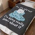 A dark gray fleece blanket with a cute cartoon cat wrapped in a blue blanket and white text reading 'yes, I'm cold but I'm being really brave about it', shown on a bed in a bedroom setting.