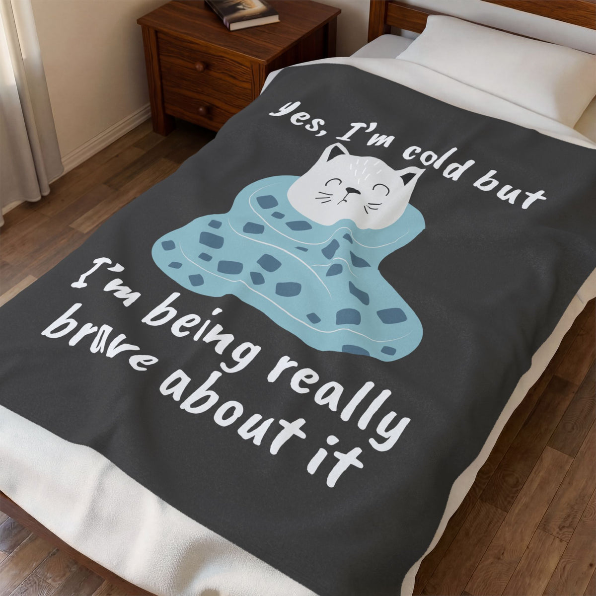 A dark gray fleece blanket with a cute cartoon cat wrapped in a blue blanket and white text reading 'yes, I'm cold but I'm being really brave about it', shown on a bed in a bedroom setting.