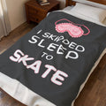 A dark gray blanket with a pink sleeping mask graphic and the text 'i skipped SLEEP to SKATE' is shown on a bed in a bedroom setting.